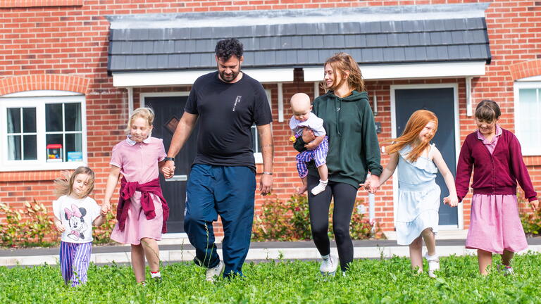 A family with two adults and five children are walking together happily on grass in front of a brick house.