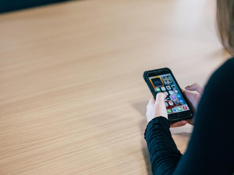A person is holding and using a smartphone while sitting at a wooden table.