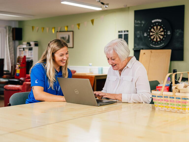 A younger woman is helping an older person use a laptop in a bright, relaxed room.