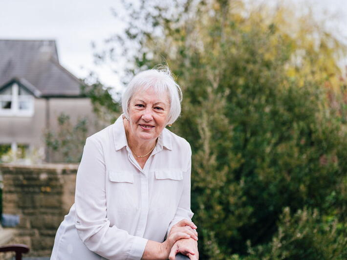 Older adult with short white hair leaning on a railing in a garden, with a house and greenery behind.
