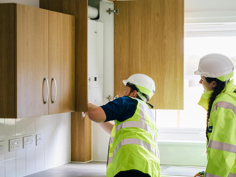 Two workers wearing hard hats and high-visibility vests are inspecting a boiler inside a kitchen cabinet.
