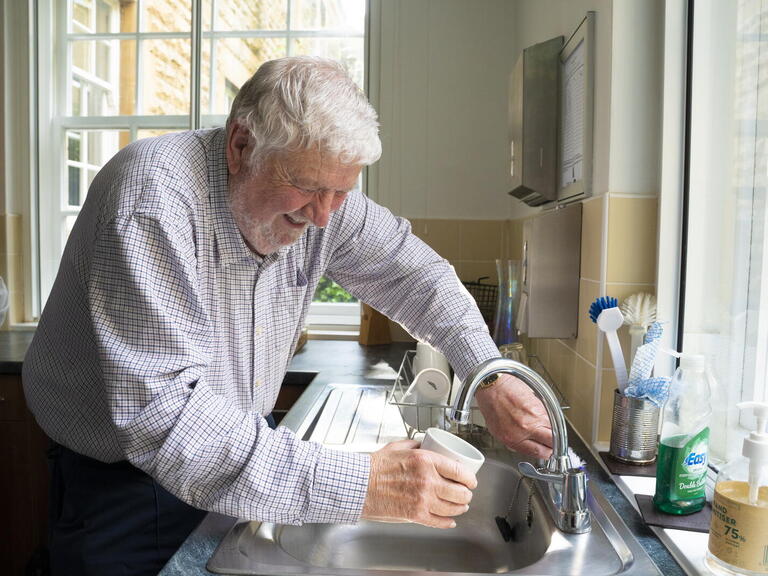 A man is filling a cup with water from a kitchen tap in a well-lit room.