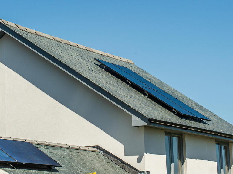 The image shows solar panels installed on the sloping roof of a white house under a clear blue sky.