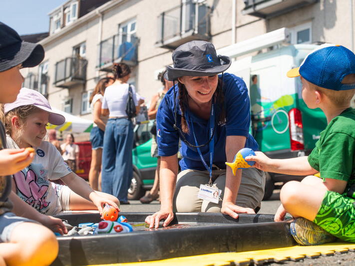 A woman in a blue shirt and sun hat plays with two children around a water tray outdoors on a sunny day.