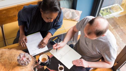 Two people sat at a table working out a budget with a calculator, pen and paper.