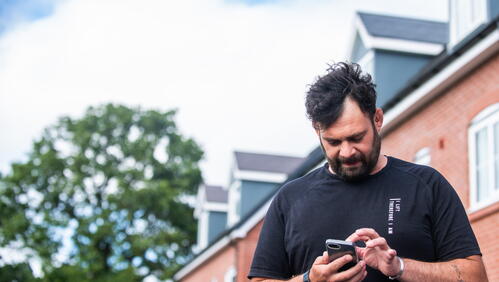 A man is using his phone outside near a row of houses on a clear day.